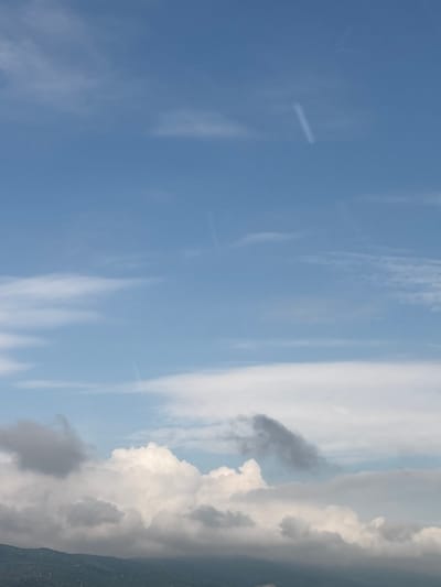 This is a vertical, outdoor, daylight photograph of a sky filled with clouds. The sky is a bright, clear blue. There are several different cloud formations visible. In the lower portion of the image, there are large, white, fluffy cumulus clouds, some of which are shaded grey, indicating depth and density. Above these, there are thinner, wispy cirrus clouds. Faint, white lines, likely contrails from aircraft, are also visible in the upper half of the sky. The bottom edge of the image shows the dark, distant silhouette of rolling hills covered in vegetation, suggesting a natural landscape beneath the sky. The scene is devoid of any human subjects or structures that would indicate a specific location within the city of Mirovo, Bulgaria.