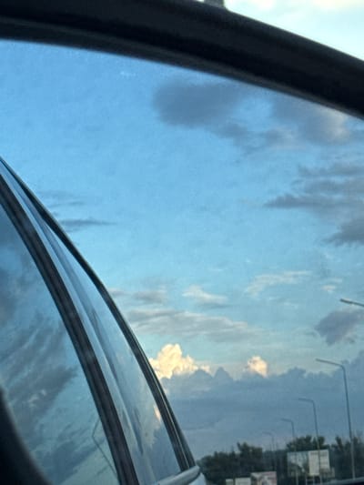 The media file shows a view through what appears to be a car window or sunroof. The primary subject is the sky, with a mix of blue sky and white, fluffy clouds. The clouds are scattered and appear to be in motion, suggesting wind. In the reflection on the glass, several light poles and trees are visible, indicating an outdoor setting, likely a roadside environment. There are also some rectangular signs visible in the reflection, though their text is illegible. The overall lighting suggests it is daytime, possibly late afternoon or early morning given the soft light and the presence of clouds. The scene conveys a sense of travel or observation from a stationary or moving vehicle. There are no people visible. The setting appears to be a somewhat rural or suburban area based on the presence of trees and light poles. The image is taken from a low angle, looking upwards towards the sky. The colors are dominated by blues and whites, with hints of green and darker tones from the reflections.