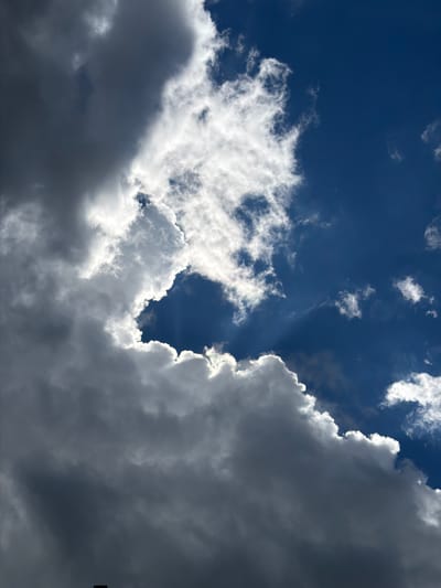 This is a close-up, vertical shot of a dramatic sky filled with dark and light clouds, set against a deep blue sky. The sun is partially obscured by thick, billowing clouds, casting bright rays through the gaps. The clouds are varied in texture and shade, with some appearing white and wispy, while others are dark and heavy, suggesting an impending storm or the remnants of one. The overall impression is one of dynamic weather and atmospheric intensity. There are no visible people, subjects, or specific objects in this image, and no recognizable landmarks that would indicate a specific city or location. It is a purely atmospheric, sky-focused photograph.