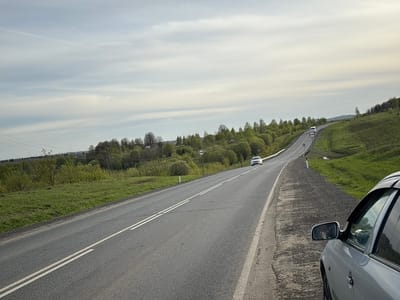 The image depicts a rural road scene near Бикбарда, Russia. The road is paved and has double solid white lines indicating no passing. Green fields and trees line both sides of the road, with some small hills in the background. The sky is overcast with various shades of gray and white clouds. There are at least two cars visible on the road, one closer and one further away. A roadside sign with a white circle and a horizontal line indicating a prohibition is present. A silver car is partially visible on the right side of the frame, with its side mirror showing a reflection. The ground immediately adjacent to the road appears to be a mix of gravel and dirt.