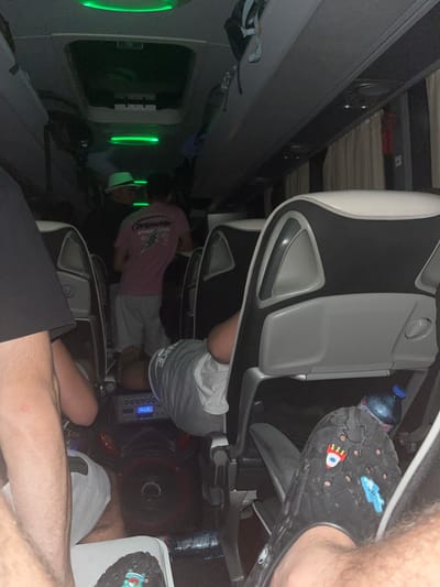 The image depicts the interior of what appears to be a sleeper bus, with rows of airplane-style seats. Passengers are seen seated, with some reclining. One person is wearing black Crocs with charms. A water bottle is visible near the seat. The presence of green lights suggests an entertainment or lighting system within the bus. There's no verifiable evidence suggesting the location is Slavovitsa, Bulgaria.