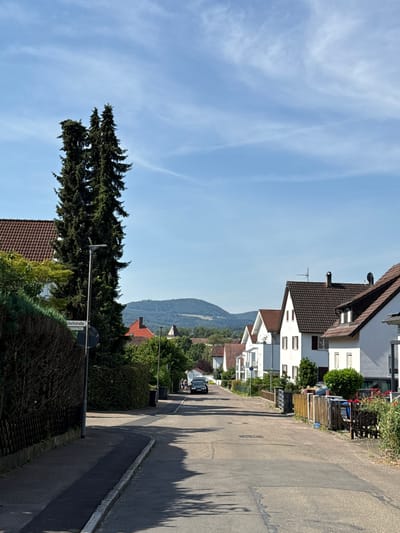 This media file depicts a street scene in Salach, Germany, on a clear, sunny day. The image is oriented vertically, with the sky occupying the top portion and the street and houses running down the right side. The scene appears to be a residential area, with several multi-story houses lining the street. These houses are primarily white with brown tiled roofs and feature multiple windows.
Vegetation is abundant, with trees and hedges flanking the street and surrounding the houses. A tall, dark green coniferous tree stands prominently on the left side of the frame, its branches reaching towards the sky. In the background, a tree-covered hill or mountain range is visible, suggesting a somewhat rural or suburban setting.
The street itself is paved and appears to be relatively quiet. Shadows from trees are cast across the asphalt, indicating the time of day is likely late morning or early afternoon, given the angle of the sun. There are a few parked cars visible further down the street.
A street sign is partially visible on the right side of the image, with the text "chottstraße" clearly legible, indicating a street name. The overall impression is one of a peaceful, well-maintained residential neighborhood in a scenic location. There are no people visible in the image. The weather is fair, with a bright blue sky and wispy clouds.