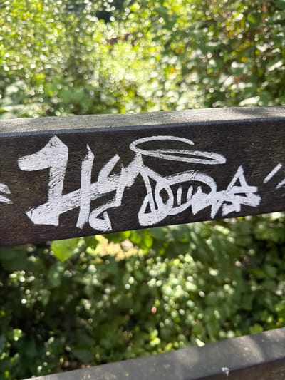 The image depicts a close-up, horizontal view of a dark, textured surface, likely a railing or beam, situated in Southend-on-Sea, United Kingdom. Applied to this surface is white graffiti, consisting of a prominent numeral "1" on the left, followed by a highly stylized character with a significant vertical stroke, and then a sequence of interconnected, angular, and curved white strokes forming a single tag extending towards the right. The paint appears to be thick and unevenly applied. The background, above and below the dark surface, is a heavily out-of-focus expanse of green foliage with numerous bright, circular bokeh light effects, indicating an outdoor, natural environment under bright, natural light. No individuals, actions, or interactions are visible.