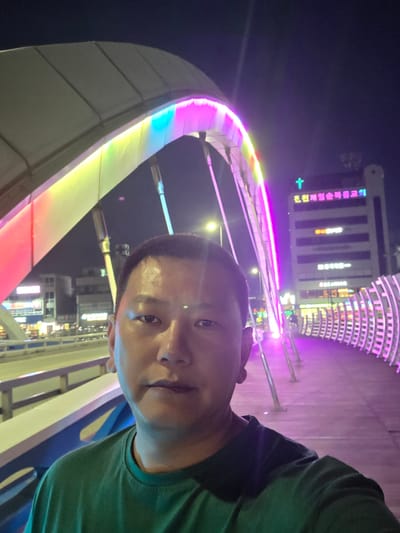 A man of Asian descent is taking a selfie at night on a pedestrian bridge in Eumnae-ri, South Korea. The bridge features an illuminated, arched canopy with vibrant pink and yellow lights. In the background, a building with Korean text and a green cross is visible, likely a church. The man is wearing a dark green t-shirt. The scene appears to be urban with city lights and structures forming the backdrop.