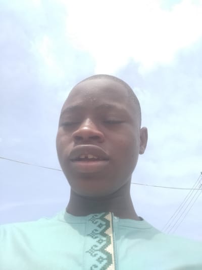 A close-up, upward-angled shot of a young man with dark skin. He is wearing a light blue shirt with a decorative patterned collar. His eyes are partially closed. Behind him, the sky is visible with some clouds. The location is likely Epe, Nigeria. Power lines can be seen in the upper right corner of the image.
