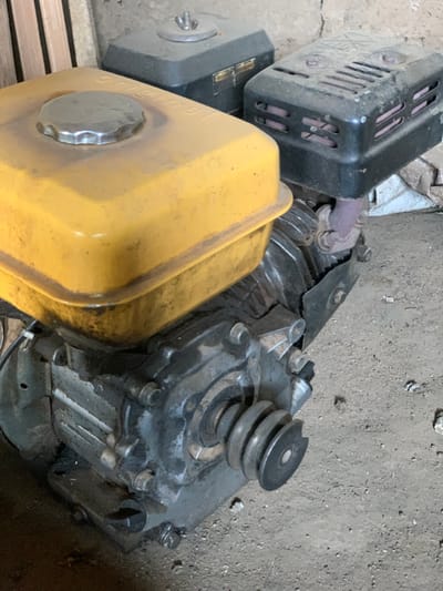 The image shows a close-up view of a small gasoline engine. The engine is a black, grey, and yellow color, and it is lying on its side on a concrete surface. The engine has a large yellow gas tank with a black cap on the top. The engine is located in the city of Egbeda, Nigeria.