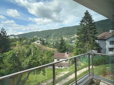 The image shows a view from a balcony overlooking a residential area with lush greenery, a lake, and a forested mountain in the background. The scene appears to be in or near the village of Kokalyane, Bulgaria, known for its natural beauty and proximity to Pancharevo Lake.
In the foreground, a modern balcony railing with glass panels and brushed metal supports frames the view. Below the balcony, a well-maintained lawn surrounds a winding stone path. Several trees, some with dense foliage and others evergreen, are scattered throughout the landscape.
Several houses are visible in the middle ground, featuring traditional tiled roofs and light-colored facades. One house, in particular, stands out with its brown roof, red brick chimney, and multiple windows. Further in the distance, a larger, white house with a flat roof is nestled amongst the trees on a hillside.
The background is dominated by a serene lake, reflecting the sky, and a vast, densely forested mountain range that stretches across the horizon. The sky is a mix of blue with scattered white clouds, suggesting fair weather. The overall impression is one of peaceful, rural living surrounded by nature.
There are no people visible in the image, and no specific actions or interactions are depicted. The focus is on the landscape and the residential architecture integrated within it.