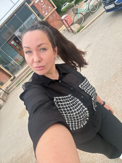 A woman with blue eyes and dark hair pulled into a ponytail is standing on an asphalt surface. She is wearing a black sheer button-up shirt with tweed-like black and white patterned pockets and black pants. The background shows a building with large windows and brick sections, a bicycle, and a blue car with a Russian license plate (H535BX 159). The scene appears to be outdoors in what could be an urban or semi-urban environment.