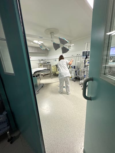 The image depicts an interior view of a medical facility, specifically a surgical or intensive care room, located in Tourcoing, France. The scene is observed through partially open, teal-colored double doors.
In the foreground, the inner frame of the left door and the partially open right door with a silver, U-shaped pull handle are visible. Both doors feature rectangular windows.
Inside the brightly lit room, a single individual, seen from behind, occupies the mid-ground. This person wears a white medical scrub top, white scrub pants, and has dark hair tied back. They are actively engaged in cleaning the speckled light-grey floor with a long-handled cleaning tool, likely a mop or squeegee, held with both hands. A small green "wet floor" or caution sign is positioned on the floor near their feet.
The room features white walls with a horizontal teal stripe at mid-height, equipped with wall-mounted medical gas outlets, electrical outlets, and rails. The white ceiling supports two large, multi-panel surgical or examination lights; one appears rectangular, and the more central one has four segmented, adjustable light panels. A grey, gridded ventilation system is visible on the upper left wall.
Medical equipment is strategically placed throughout the room. On the left, a gurney or operating table with white linens is present, alongside a stainless steel medical cart. Various other metallic stands and equipment are also visible. On the right, a complex setup of medical monitoring devices, including at least two screens displaying graphical information, is visible, accompanied by an array of tubing, wires, and other clinical apparatus. The overall environment appears meticulously clean and organized.