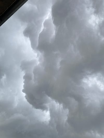 A low-angle view captures a sky filled with ominous, dark grey storm clouds. The clouds are dense and turbulent, suggesting an impending weather event. In the upper left corner, a portion of a dark, textured surface, possibly the underside of a roof or overhang, juts into the frame, providing a stark contrast to the vast expanse of cloud. No people or specific landmarks are visible in this image, which focuses solely on the atmospheric conditions. The location is identified as Uchaly, Russia.