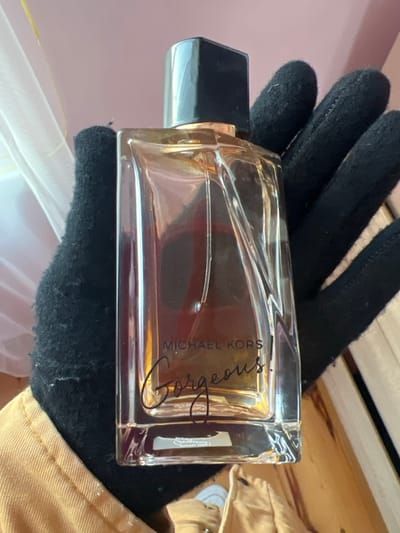 A hand in a black glove holds a bottle of Michael Kors "Gorgeous" perfume. The bottle is rectangular with a clear body and a black cap. The perfume liquid inside is a brownish-amber color. The label features the brand name "MICHAEL KORS" in smaller letters above the word "Gorgeous" in a larger, script-like font. Part of a light-colored jacket is visible on the right. The background includes a hint of pink wall and wooden planks.