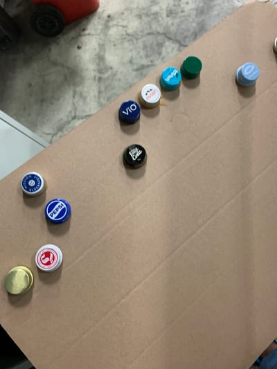 A collection of bottle caps are arranged on a piece of cardboard. The cardboard is placed on a concrete surface. In the background, a red object with a black wheel is visible. The bottle caps include brands such as Vio, Evian, Vita Cola, Pepsi, and two others that are not clearly identifiable. One cap is gold colored and appears to be made of metal. The scene appears to be indoors or under cover, as indicated by the smooth, grey concrete floor. There is no indication of the specific location within Worms, Germany.
