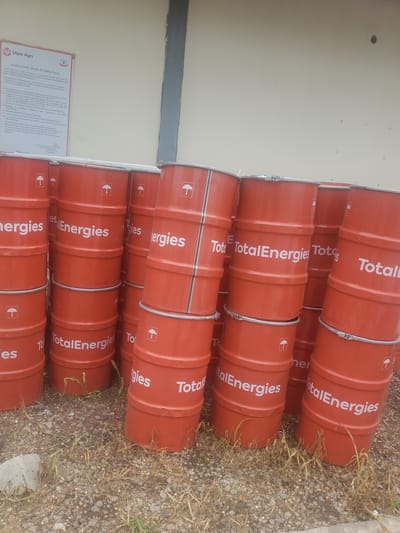 In the city of Gwagwada, Nigeria, a stack of bright orange, cylindrical barrels with white "TotalEnergies" branding and umbrella logos are arranged in uneven piles against a light-colored wall. The barrels are stacked several high, with some leaning precariously. Dry grass and gravel cover the ground around the barrels. A portion of a document with "Olam Agri" and "Environment, Health & Safety Policy" visible is also present in the upper left corner of the image.