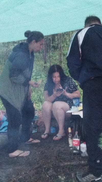 In Tarasovskoe, Russia, three individuals are present under a light blue tarp shelter. One person, on the left, stands wearing a dark jacket and dark pants. Another individual, seated in the center, wears a black and white patterned shirt and is looking at a mobile phone. To the right, a third person, wearing a dark jacket with white accents, stands with their back to the camera. The ground beneath them is covered in grass and dirt. On a white surface to the right of the seated person are two bottles, one of which is a Coca-Cola bottle. The environment appears to be wooded with trees visible in the background.