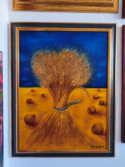 A framed painting of wheat stalks tied with a blue ribbon hangs on a white wall in Kerpen, Germany. The painting depicts a large bundle of golden wheat against a deep blue sky, with a field of yellow below. Several small, round hay bales are scattered across the field. The artist's signature, "Amanda Anna," is visible in the bottom right corner of the painting. The painting is framed by a dark blue border and a thin gold inner frame. Another framed painting with a gold frame is visible above it.
