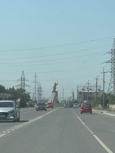 The media file depicts a view from a vehicle traveling on a highway in Ынтымак, Kyrgyzstan. The scene is dominated by a clear, pale blue sky and a long, straight asphalt road with white lane markings. Tall electrical pylons line the left side of the road, supporting numerous power lines that stretch across the frame. In the distance, on the left, a golden statue of a figure holding a spear is visible atop a pedestal, framed by the electrical infrastructure.

Several cars are on the road, suggesting traffic is flowing. A red car is prominent in the middle distance, followed by a dark-colored car and then a silver SUV in the foreground. The sun appears to be high in the sky, indicating it is daytime, likely midday, with fair weather. The presence of modern vehicles and the highway infrastructure suggests a developed or developing area within Kyrgyzstan. There is no indication of people in detail, but the cars imply their presence. The overall mood is neutral, capturing a common travel scene. Visible text on the license plate of the silver SUV reads "371 APX".