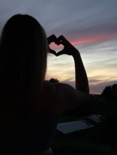 A silhouetted individual with blonde hair is facing away from the camera, forming a heart shape with their hands in front of a twilight sky. The sky is streaked with hues of purple, pink, and yellow, indicating either sunrise or sunset. Below the sky, a dark silhouette of trees and foliage is visible, with two small greenhouses or garden structures appearing in the lower right portion of the image. The location is identified as Dreiliņi, Latvia.