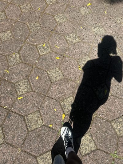The image presents a top-down view of paved ground, featuring a prominent, elongated shadow of a person and their feet. The shadow occupies a significant portion of the frame, stretching from the upper left towards the lower right, indicating a strong light source, likely the sun, positioned at an angle.
In the lower right corner, the person's feet are visible, wearing white athletic shoes with black laces and dark socks. The shoes appear to be modern, possibly running or walking shoes, with visible mesh or perforated designs.
The ground is composed of textured paving stones arranged in a geometric pattern of alternating octagonal and square blocks. The stones are predominantly grey and light brown hues, with some areas appearing lighter or darker due to variations in material or wear. Scattered across the paved surface are several small, dried yellow leaves, suggesting an outdoor environment, possibly a sidewalk or courtyard.
The stark contrast between the deep black shadow and the brightly lit pavement indicates a clear, sunny day. The length and direction of the shadow suggest the time of day is likely morning or late afternoon. No specific activity beyond standing or walking is depicted. There is no visible text within the image. The scene is set in Santa Tecla, El Salvador.