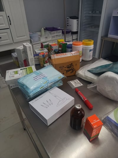 The image shows a cluttered stainless steel table in what appears to be a medical or veterinary setting. A variety of medical supplies, including surgical gauze in sterile packaging, suture materials ("MAX Chromic Catgut"), medications like "FENAM" and "MEIXYLA," and a device labeled "Gutta Cutter," are scattered across the surface. The presence of a "Thank you" box suggests a transactional aspect, possibly related to the acquisition or distribution of the supplies. In the background are white cabinets and a refrigerator. The text on some of the packaging suggests the location of Thong Sala, Thailand.
