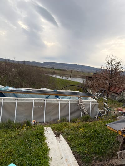 The image shows an above-ground Intex swimming pool set up in an outdoor area, likely a yard or garden, in Liaskovo, Bulgaria. The pool is gray and white, with visible metal support posts. A ladder leans against the side of the pool. The pool appears to be partially covered. A multi-colored beach ball is near the base of the pool.

The surrounding environment includes green grass and other vegetation. Debris and miscellaneous objects are scattered in the area, indicating a state of disarray. In the background, a rural landscape extends toward the horizon, including a body of water (possibly a lake or river), a building with a red tile roof, hills, and bare trees. The sky is overcast.