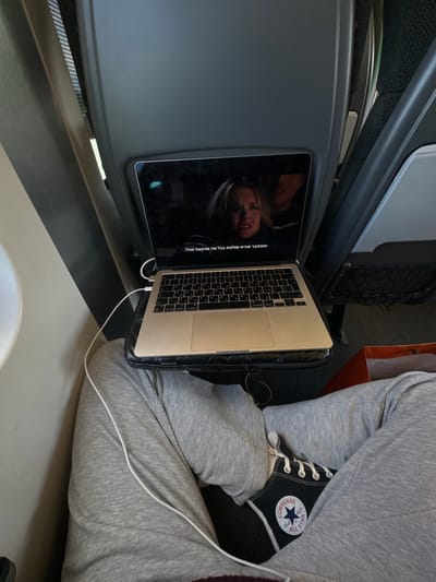 A person is sitting in a train seat, wearing grey sweatpants and black Converse sneakers. They are using a laptop computer to watch a movie on a dark screen. The laptop is resting on a black tray table that is positioned over the person's lap. The movie features a woman with long dark hair who is looking at the camera with a serious expression. The image was taken in the city of Enns, Austria at the coordinates 48.227 latitude and 14.468 longitude.  A white cable is connected to the laptop. A red shopping bag is visible on the right side of the image.  The train seat is grey and has a dark headrest. The image appears to be taken from an overhead angle.  The ambient light is dim.