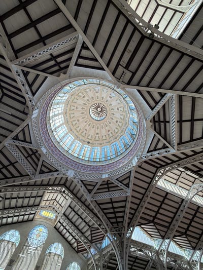 The image shows an upward view of the interior ceiling structure of the Central Market of Valencia, Spain. The architecture is characterized by an ornate, circular glass dome at the apex, decorated with floral motifs and geometric patterns. This dome is surrounded by a complex system of white metal trusses and beams, which support a sloped roof structure made of wooden planks and glass panels, allowing natural light to filter in. Below the central dome, the market's main hall features arched stained-glass windows with heraldic designs, adding color and intricate detail to the scene. The overall impression is one of grand scale and historical architectural style, with the interplay of light and shadow highlighting the intricate details of the market's construction.