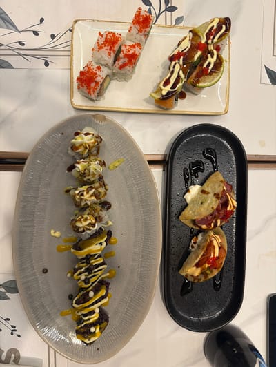 A top-down shot shows an assortment of sushi and sushi-like items arranged on white and gray oval plates and a black rectangular plate, all set on a white marble-patterned surface. In the upper left, a white rectangular plate holds several pieces of sushi rolled in rice and topped with bright orange roe. To the right of this, a similar plate features sushi rolls garnished with thin slices of lime, drizzled with white sauce and a dark, glossy sauce, and sprinkled with more orange roe. Below these, on a large, gray, oval-shaped plate, a vertical arrangement of six sushi rolls is topped with a creamy white sauce, a dark drizzle, and accents of yellow sauce and dark sprinkles. On the right side, a black rectangular plate holds two items that resemble tacos, filled with what appears to be raw fish, cream sauce, and red roe, and also drizzled with a dark sauce. The background features a subtle decorative pattern of gray leaves and branches on a white surface. No people are visible in the image, and the scene appears to be an indoor food setting, possibly a restaurant. The city of Ondara, Spain, is not depicted or referenced in the image.