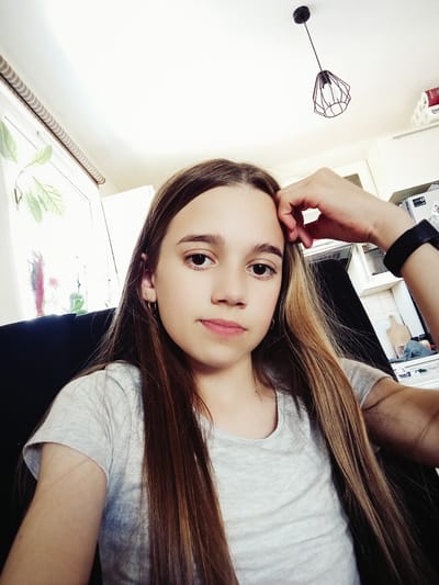 The media file is a selfie of a young person with long, brown hair. The person is wearing a light grey t-shirt and has their right hand resting on their face. Their expression is neutral, and the lighting suggests it's daytime, possibly indoors near a window. In the background, a white wall is visible with a modern black light fixture. There are also glimpses of what appears to be a kitchen or room with some appliances and shelving units. A roll of toilet paper with some visible red text is also in the background. The image is slightly angled.
