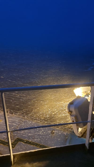 A ship's railing is in the foreground of the image, with a bright light illuminating falling snow or sleet. The ocean is visible in the background, with the dark blue sky above. The scene suggests a cold, maritime environment, possibly at night or during heavy snowfall. There are no visible people or buildings. The location is unknown.