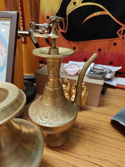 The image shows a close-up of an ornate brass hookah with intricate carvings, placed on a wooden surface. The hookah is the primary subject, with a falcon-shaped handle at the top and a curved spout. To the left of the hookah, the edge of a framed picture and a yellow cylindrical object with a silver tap are visible. In the background, a cardboard box, some papers, and a tapestry with a star pattern can be seen. The scene appears to be indoors, possibly in a shop or a home in Kathmandu Metropolitan City, Nepal, given the cultural aesthetic of the hookah and the tapestry. There are no people present in the image.