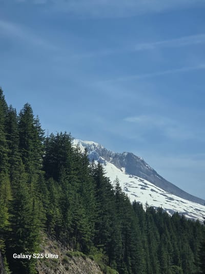 The image shows a snow-capped mountain peak with a forest of evergreen trees in the foreground. The sky is clear and blue with some wispy clouds. The text "Galaxy S25 Ultra" is visible in the lower left corner of the image. This scene is consistent with views of Mount Hood from Government Camp, Oregon, USA.