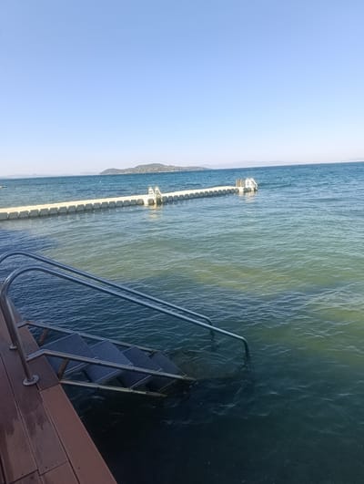 The image depicts a serene coastal scene in Urla, Turkey, under a clear, bright sky, suggesting it's daytime with good weather. The foreground features a wooden deck on the left, with a set of metal stairs leading down into the clear blue water. The stairs are partially submerged, indicating the water level is high or the tide is in. The water itself is calm, with gentle ripples creating subtle patterns on its surface.

In the middle ground, a modular floating dock stretches horizontally across the water. This dock is composed of numerous interconnected rectangular modules, creating a stable platform on the sea. At the far end of the dock, another set of stairs, similar to the one in the foreground, descends into the water, complete with handrails.

In the background, the horizon is marked by a distant landmass, likely an island or coastline, which appears green and hilly. The sky is a vast expanse of uninterrupted blue, free of clouds. The overall impression is one of tranquility and a peaceful day by the sea, perhaps a location for swimming or water activities. There is no visible text in the image.