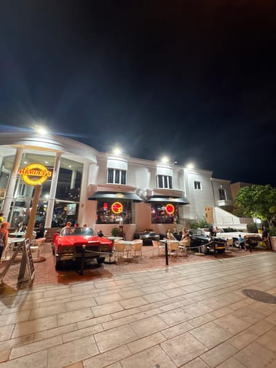 The image depicts an outdoor scene at night in Adeje, Spain, in front of a building with a bright, illuminated facade. The building appears to be a restaurant or bar named "AMERICAN HARLEYS," as indicated by a large neon sign.
Several people are visible in the scene. Some are seated at tables outside the establishment, enjoying what appears to be a meal or drinks. Others are inside the building, which is also lit and seems to have patrons. A striking red vintage car is parked on the pedestrian walkway, possibly as a decorative element or a photo opportunity for customers. The walkway is paved with rectangular stone tiles and is illuminated by the building's lights and some streetlights.
The time of day is clearly night, with the dark sky providing a contrast to the bright lights of the building and the neon sign. The weather appears to be clear, as there are no indications of rain or other adverse conditions. The overall atmosphere seems to be lively and social, with people gathered and enjoying the evening. The presence of outdoor seating and dining suggests a pleasant climate suitable for al fresco activities.
Visible text includes the prominent "AMERICAN HARLEYS" sign, which is circular with neon lettering. Other signs with text are partially visible inside the establishment. The scene suggests a popular spot for evening entertainment and dining in Adeje.