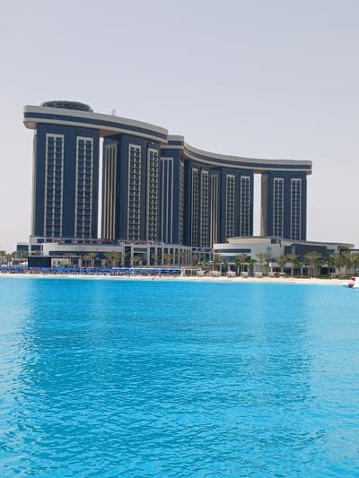The image shows the Rixos Alamein hotel in Sidi Abdel Rahman, Egypt, alongside a large turquoise-colored lagoon. The hotel is a tall, multi-story building with a distinctive architectural design featuring stacked, horizontal sections. Palm trees and beach umbrellas line the edge of a sandy area between the hotel and the water. The sky is a pale color, suggesting clear or hazy weather.