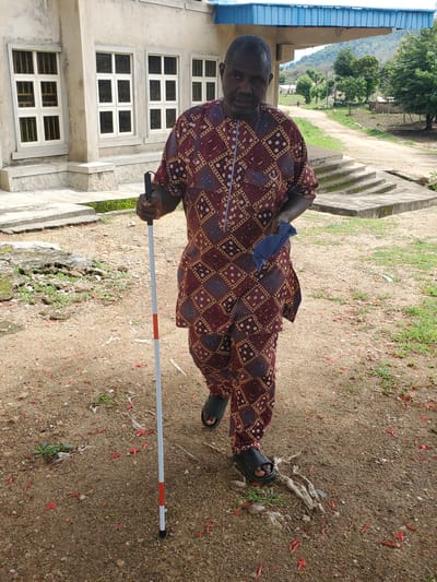 A man stands outdoors near a building, holding a white and red tipped cane. He is wearing a maroon and gold patterned traditional African outfit and black sandals. The man appears to be partially blind or visually impaired and is using the cane to navigate his surroundings.

The setting is outdoors, with a building that has multiple windows on the left side of the frame. The ground is covered in dirt, pebbles, and some green vegetation, with scattered red petals on the ground. In the background, there are trees and a hillside. The lighting suggests it is daytime. The image is taken from a slightly elevated, rotated perspective. The overall mood is neutral, capturing a moment of the man in his environment. There is no visible text in the image.