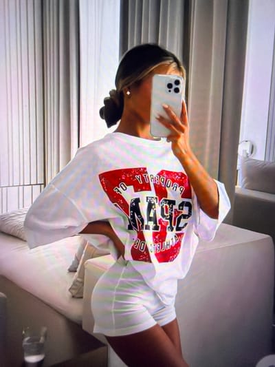 A young woman with her hair in a bun is taking a mirror selfie in what appears to be a bedroom. She is wearing an oversized white t-shirt with a graphic print on the back that includes red lettering and the numbers "492". She is also wearing white shorts. The room has light-colored vertical blinds behind her, and a light-colored sofa or bed is to her left. A glass with a light-colored liquid is on a surface in the foreground.