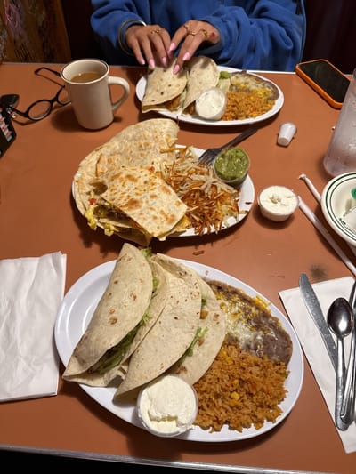A table is set with plates of Mexican food, including tacos, quesadillas, rice, beans, hash browns, guacamole, and sour cream. A person is seated, their hands visible with multiple rings. There is also a mug of coffee, glasses, a cell phone, and utensils. The location is Huntington Beach, United States.