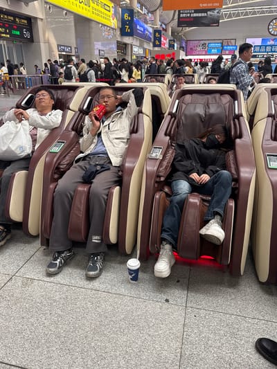 The image shows a row of massage chairs occupied by people in what appears to be a waiting area, likely in a public transport hub like a train station or airport. The chairs are dark brown with beige accents, and several people are seated in them, some appearing to be resting or sleeping. One person is holding a red package. The area is crowded with many other people visible in the background, and signs with text and numbers are hanging from the ceiling. The floor is made of gray speckled tiles. The photo was taken indoors.

Based on the signs including characters, this photo likely was taken in Shenhe District, China.