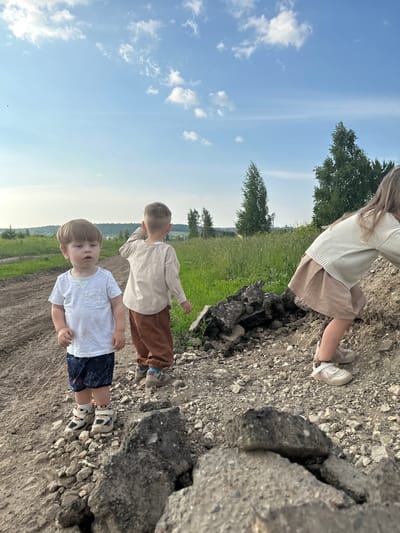 Three children are on a dirt path near a grassy field under a partly cloudy blue sky. In the foreground to the left, a young boy with light brown hair is looking slightly to his left. He is wearing a white t-shirt, dark blue shorts with a white pattern, and light-colored sneakers. Behind him and to the right, another young boy with light brown hair is facing away from the camera, looking towards the right. He is wearing a light beige long-sleeved shirt and brown pants, with light-colored sneakers. To the far right, a child with long blonde hair is leaning forward and looking down, partially obscured by a pile of rocks. This child is wearing a light beige sweater and a light brown skirt, with light-colored sneakers. The environment appears rural, with tall grass and trees visible in the background. The path is composed of dirt and gravel, with scattered rocks and some broken asphalt pieces in the foreground. There is no indication of the city of Estachi, Russia in the image.