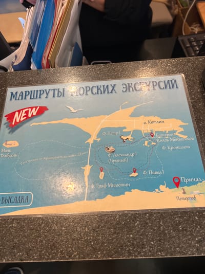 The image shows a map with a title in Russian that translates to "Sea Excursion Routes". The map depicts various islands, forts, and locations, likely in the vicinity of Kronstadt, Russia. Key locations marked include "Fort Peter I", "Fort Alexander I (Chumny)", "Fort Menshikov", "Fort Kronshlot", "Cape Kotlin", "Graf Million", "Tolbukhin Lighthouse", and "Pier Peterhof". A red marker indicates "Pier" (Причал). The map also shows a dotted line indicating a route, with a label mentioning "Distance from Kronstadt to lighthouse 2.8 miles". There is a red "NEW" tag on the top left corner of the map. The map is placed on a dark, speckled countertop.