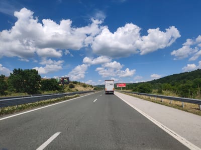 This image depicts a view from a vehicle traveling on a highway through a hilly, rural landscape. The road is asphalt with a white lane marker and a concrete shoulder, bordered by a metal guardrail. Small white flowers grow along the edge of the road next to the guardrail. On the left side of the highway, a billboard with a picture of what appears to be a food item and the word "Nuti" is visible. Further down the road, on the right side, another billboard with red lettering is present.
A white truck with "Josef Emerswieser" and other text in black lettering on its rear is in front of the viewer, traveling in the same direction. The surrounding scenery consists of green trees and dry, grassy hills. The sky is a vibrant blue with numerous fluffy white clouds, suggesting a sunny day with scattered cloud cover. The time of day appears to be midday, given the bright sunlight. The location is identified as Ravnishte, Bulgaria. No people are clearly visible in the image, and the overall mood is one of travel and open road.