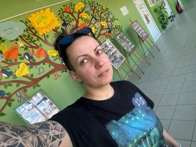 The image shows a woman in an indoor space, likely a school or community center, in Radomir, Bulgaria, with a wall decorated with a large tree mural. The mural features a tree with owls, leaves, and labels in Bulgarian, promoting values like "friendship," "respect," "sincerity," "gratitude," and "tolerance." She is wearing a black shirt with an inscription that reads "1883."  She is also wearing sunglasses and has tattoos on her right arm. The walls are green, and framed photos are displayed. The floor is tiled in neutral tones.