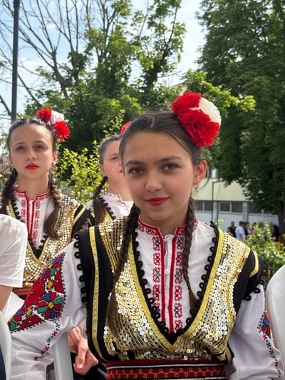 The image shows three young girls in traditional Bulgarian folk costumes in Pordim, Bulgaria. The girl in the foreground, on the right, is looking directly at the camera with a slight smile. She has dark hair braided in pigtails and wears a red and white floral decoration in her hair. Her costume is white with intricate red embroidery on the neckline and sleeves, and is adorned with numerous gold sequins on the vest. The girl to her left, in the middle, is also looking forward, her face slightly blurred. She has similar braided hair and a red and white floral accessory. The girl on the far left is looking to her left, with her face partially obscured. She also has braided hair and a floral hairpiece. All three girls are wearing white blouses with embroidered details and elaborate vests. The background shows greenery and trees, suggesting an outdoor setting, possibly a festival or cultural event. In the further background, there are people and a building.