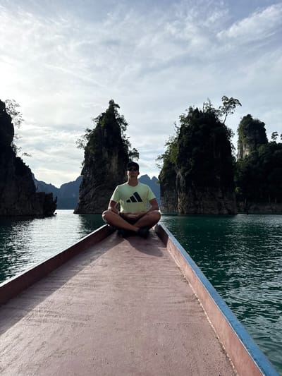In Khao Phang, Thailand, a person is seated in a lotus position on the bow of a boat, which is navigating through a body of water surrounded by dramatic limestone karsts. The individual, wearing a light-colored t-shirt and shorts, is facing the camera with eyes closed, seemingly meditating or in a state of relaxation. The boat's deck, a reddish-brown surface, occupies the foreground. The water, a deep green, reflects the overcast sky and the surrounding rock formations. The karsts, covered with lush vegetation, rise steeply from the water, creating a serene and majestic landscape. The sky is a mix of pale blue and scattered white clouds, suggesting a daytime scene.
