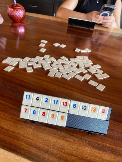 A wooden table is strewn with white game tiles, some arranged randomly, others in a holder. A person partially visible on the left holds a smartphone, possibly taking a photo of the game setup. A hexagonal object, possibly a decoration, is also present on the left edge of the table. The focus of the image is on the game tiles and their arrangement.
