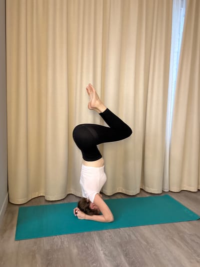 A woman is performing a yoga pose on a teal mat. She is in a headstand, with her legs bent at the knees and her feet pointed upwards. She is wearing a white tank top and black leggings. The background is a beige curtain. The floor is made of wooden planks. There is a small glimpse of a window to the right, covered by a sheer white curtain. The scene appears to be indoors, possibly in a yoga studio or a home.