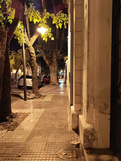 The image depicts a street scene at night in Victoria, Argentina. The sidewalk, paved with patterned tiles, is illuminated by streetlights casting a warm glow. A row of trees lines the street, their foliage partially obscuring the light. Two cars are parked along the curb - a silver car closer to the viewer, and a red car further down the street. On the right side of the frame, a building with a textured facade stands close to the sidewalk. The overall impression is a quiet, dimly lit urban street.