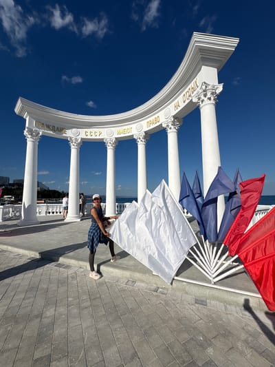 The image shows a woman standing in front of a white, classical-style archway with columns. The archway has text written on it in gold letters. To the right of the woman are several flags, some white, some blue, and some red, arranged in a fan shape. The woman is wearing a blue plaid shirt, dark leggings, and white sandals. She is holding a large white flag. In the background, there is a body of water and buildings. The sky is blue with some white clouds. The text on the archway is in Cyrillic script and reads "ГРАЖДАНЕ СССР ИМЕЮТ ПРАВО НА ОТДЫХ", which translates to "Citizens of the USSR have the right to rest." This monument is located in the city of Alushta, Ukraine.