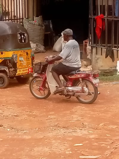 The image shows a man riding a red and white scooter in Umuokpu, Nigeria. He is wearing a light gray shirt with the word "STAFF" printed on the back, black shorts, and a gray hat. He is wearing brown sandals. There is a yellow tuk-tuk parked to the left of the scooter. The ground is red dirt. In the background, there is a building with a metal gate and bags of some sort. A red piece of cloth is draped over the gate.