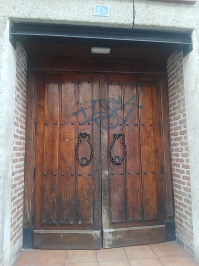 The image shows a pair of heavy, weathered wooden double doors with vertical paneling and decorative metal studs. Each door features a distinctive circular metal handle made of chain links. The doors are set within a brick facade, flanked by rough-textured concrete. Above the doors, a dark metal lintel is visible, and a light fixture is mounted on the underside of the lintel. A blue rectangular plaque with the number "25" is affixed to the concrete above the lintel. The doors are marked with black graffiti. The ground in front of the doors is paved with terracotta tiles. The overall impression is of an old, possibly rural, entrance. This is the entrance to a house in Navaluenga, Spain.